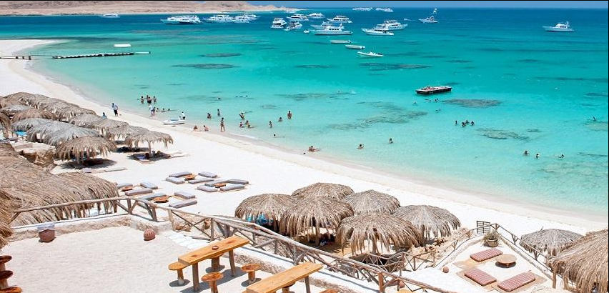 A scenic Hurghada beach overlooking the turquoise waters of the Red Sea, with boats offshore and a relaxing sandy shoreline. Hurghada is a popular destination for snorkeling, diving, island trips and beach holidays.